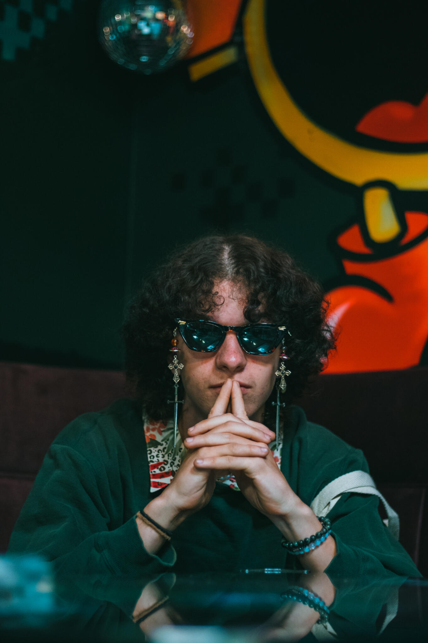 Hemp City A man with curly hair and sunglasses is sitting at a table, exuding a laid-back vibe in a social environment.